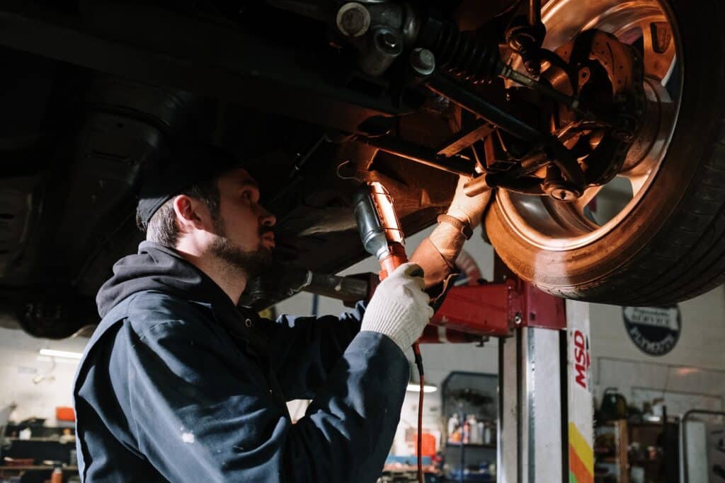 a mechanic with a beard and dark blue coveralls shines a shop light on the wheel of a car from underneath